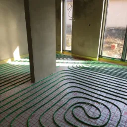 Wnętrze w trakcie montażu ogrzewania podłogowego: zielone rurki ułożone na folii z czerwoną siatką, widoczne okna i fragmenty ścian w surowym stanie.