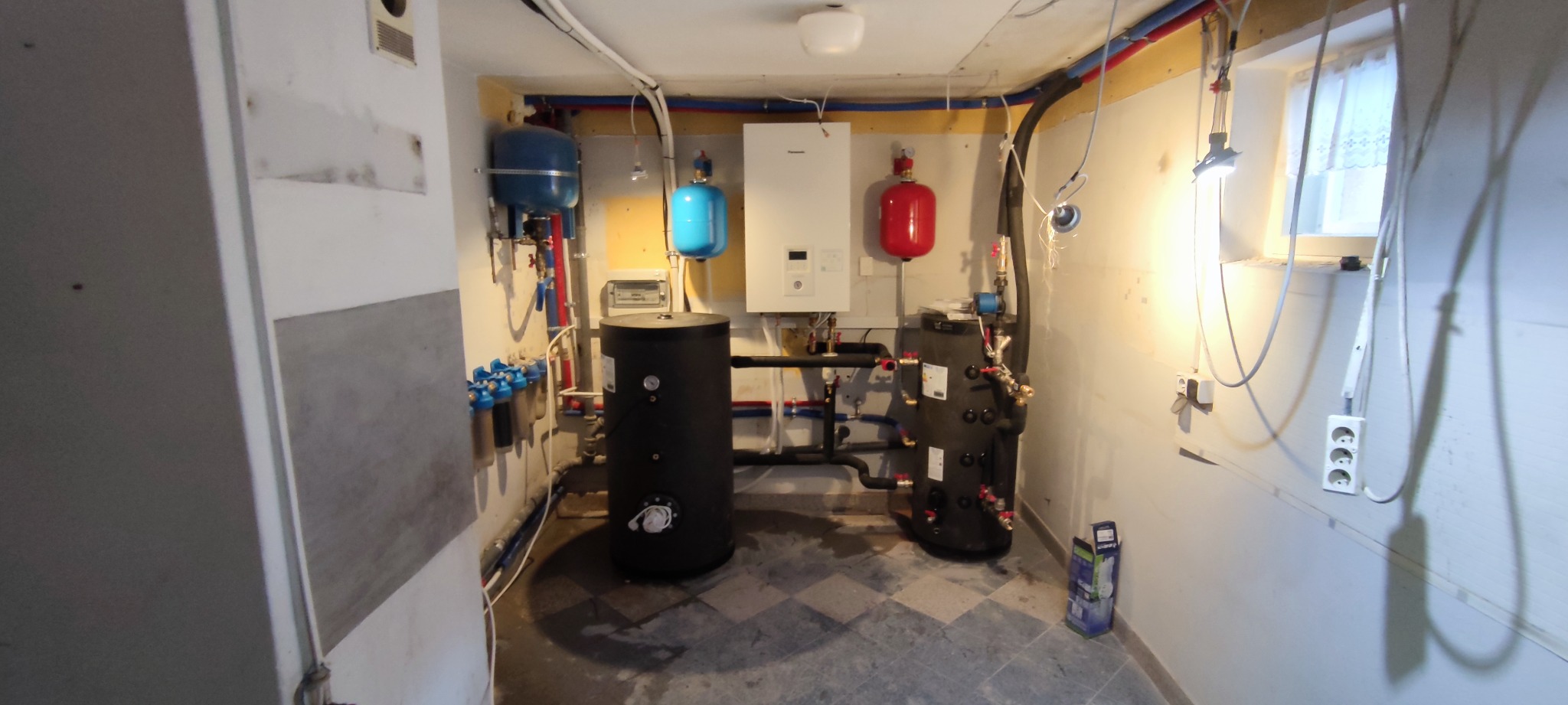 Kompleksowa instalacja CO z piecem gazowym, zbiornikami i filtrami w pomieszczeniu technicznym. Widoczne rury, zawory i okno. Szachownica płytek na podłodze.