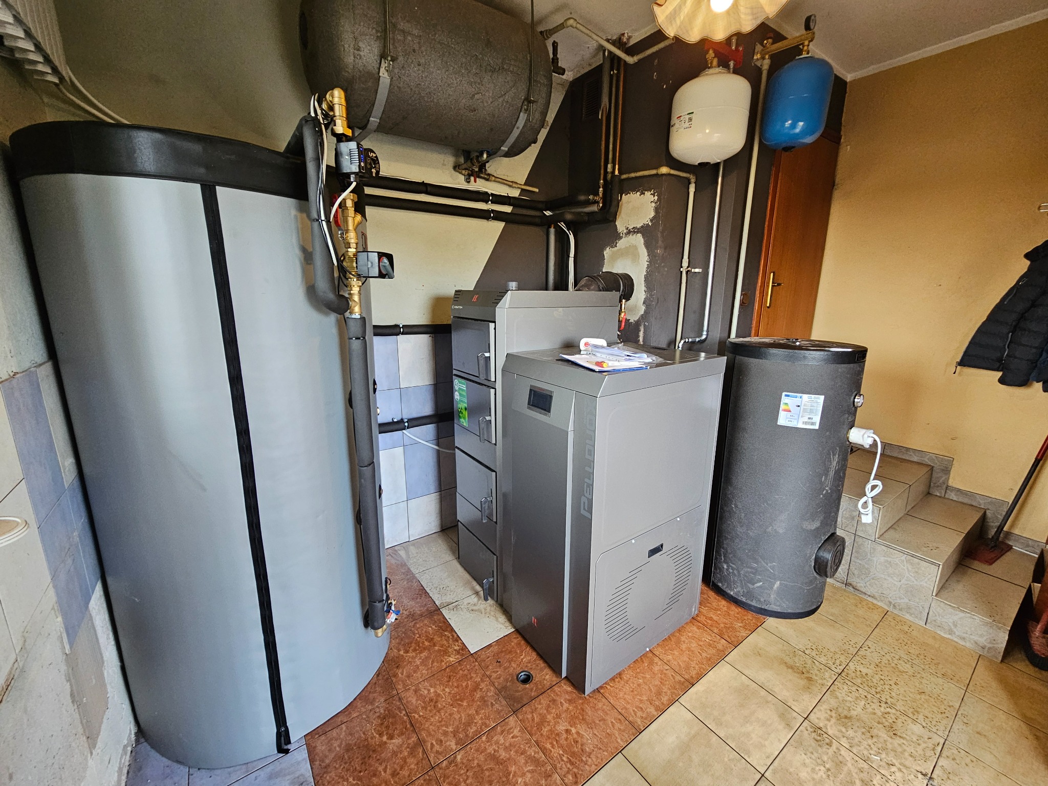 Kotłownia z nowoczesnym piecem na pellet, zbiornikiem buforowym i instalacją. Widoczne elementy instalacji grzewczej i rury. Ujęcie z perspektywy podłogi.