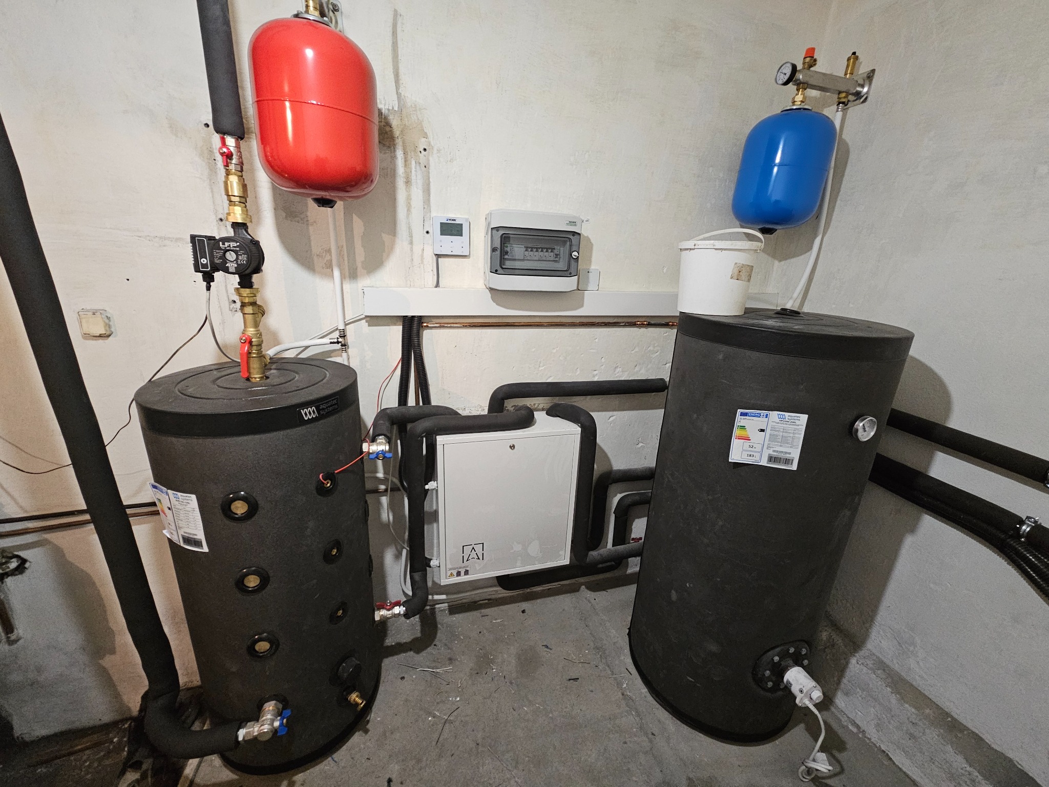 Kompletna instalacja CO z dwoma zbiornikami akumulacyjnymi, pompą obiegową i naczyniami wzbiorczymi w kotłowni. Widoczna tablica elektryczna i rury.