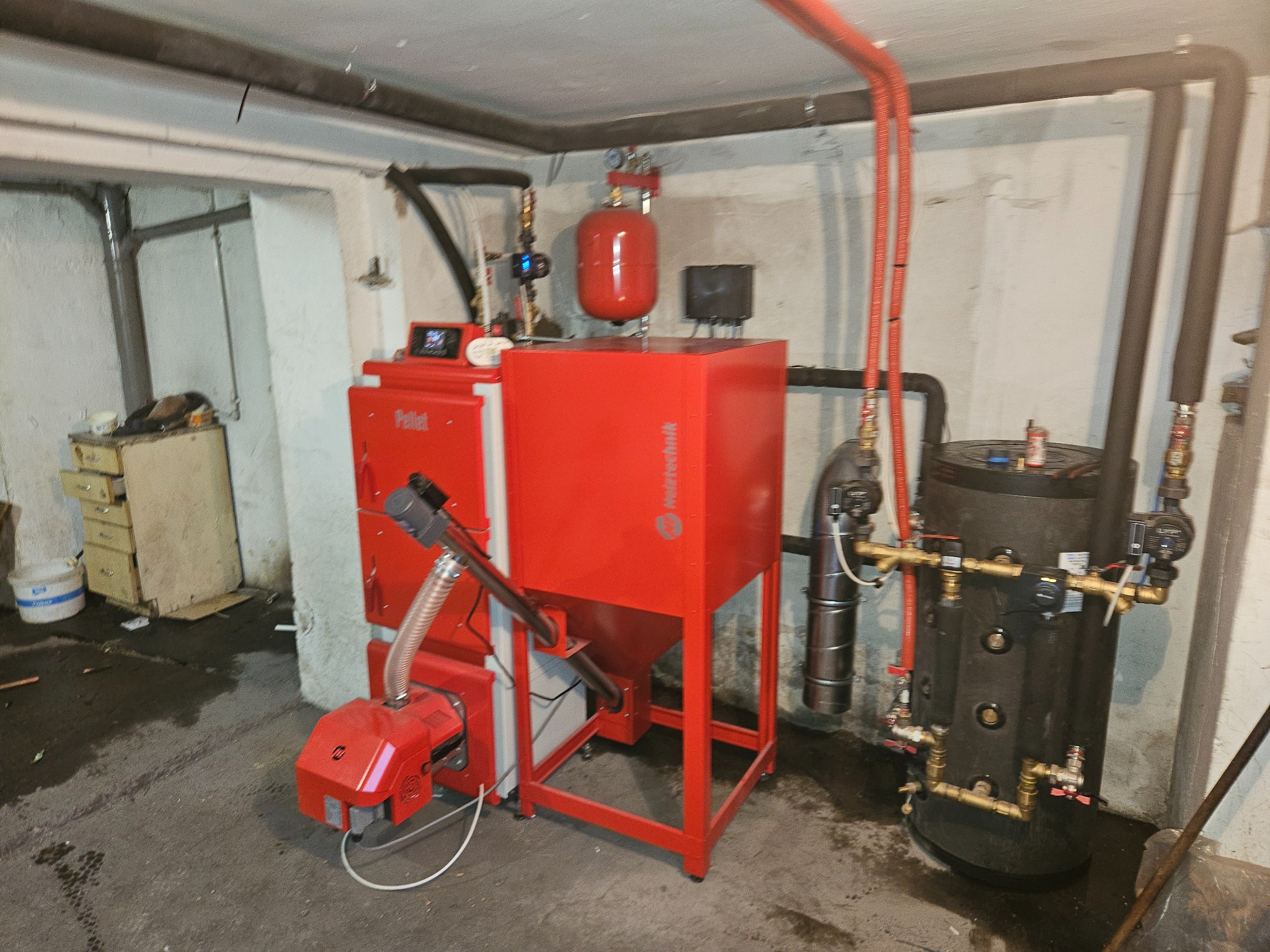 Czerwony piec na pellet Techmilk z zbiornikiem i rurami w piwnicy z widocznym osprzętem, po instalacji. Szare ściany i podłoga.