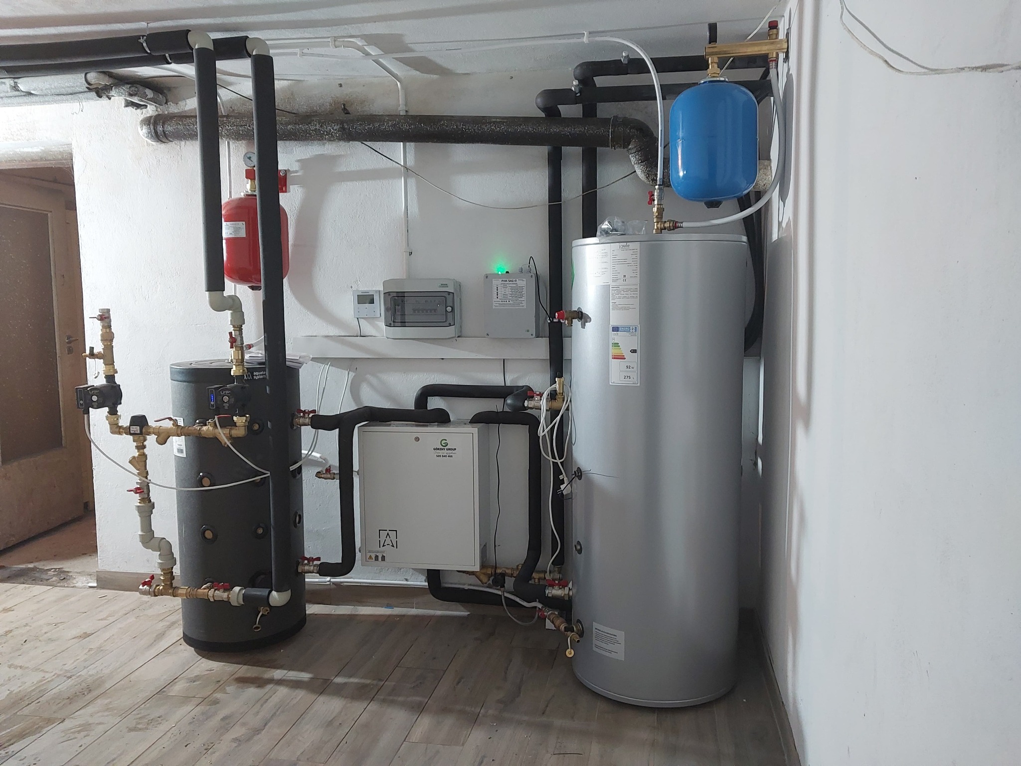 Nowoczesna kotłownia z szarym zbiornikiem i białą pompą ciepła, rury w izolacji, manometry i zawory. Instalacja w piwnicy na drewnopodobnej podłodze.