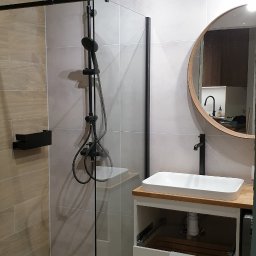 Pan Łazienka - Nowoczesna łazienka z kabiną prysznicową typu walk-in z czarnymi elementami, okrągłym lustrem z drewnianą ramą i białą umywalką nablatową na szafce z drewnianym blatem.