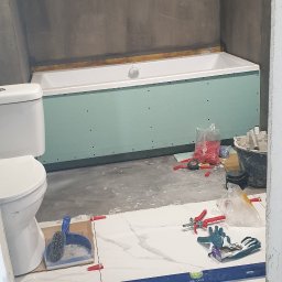 Pan Łazienka - Białe płytki podłogowe w trakcie układania w łazience, widoczny poziomica LUX-TOOLS, krzyżyki dystansowe, obudowana wanna płytą kartonowo-gipsową, oraz surowa ściana w tle.