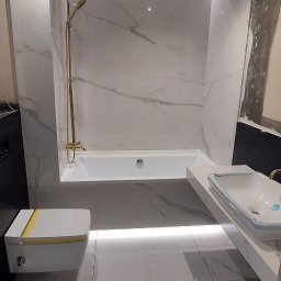 Pan Łazienka - Nowoczesna łazienka z białymi marmurowymi płytkami, złotą armaturą, podświetlaną wanną i umywalką oraz wiszącą toaletą z żółtym akcentem.