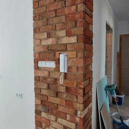 Pan Łazienka - Wykończony narożnik ściany z cegły klinkierowej wewnątrz pomieszczenia, z widocznymi gniazdkami elektrycznymi i domofonem, na tle surowej podłogi i narzędzi budowlanych.
