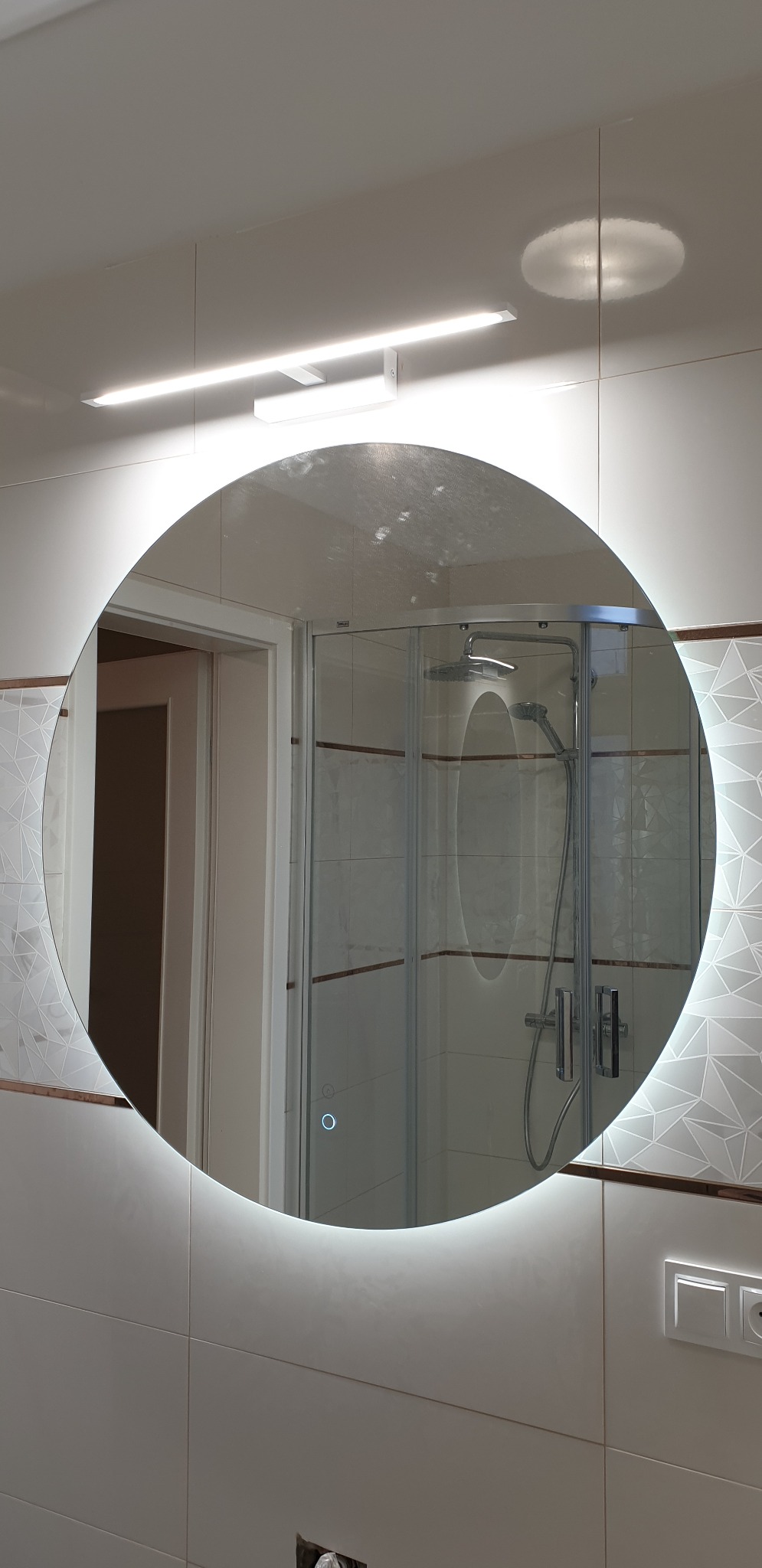 Okrągłe lustro z podświetleniem LED w wykończonej łazience, odbijające kabinę prysznicową i fragment otwartych drzwi, z widocznymi płytkami w geometryczny wzór i listwą o miedzianym odcieniu.