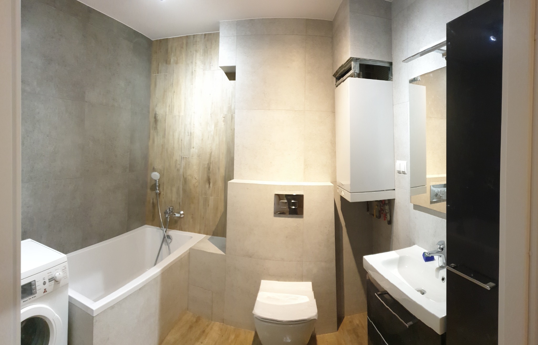 Nowoczesna łazienka z wanną, umywalką na szafce w kolorze czarnym, podwieszaną toaletą i pralką, wykończona płytkami w odcieniach szarości i beżu oraz piecem gazowym