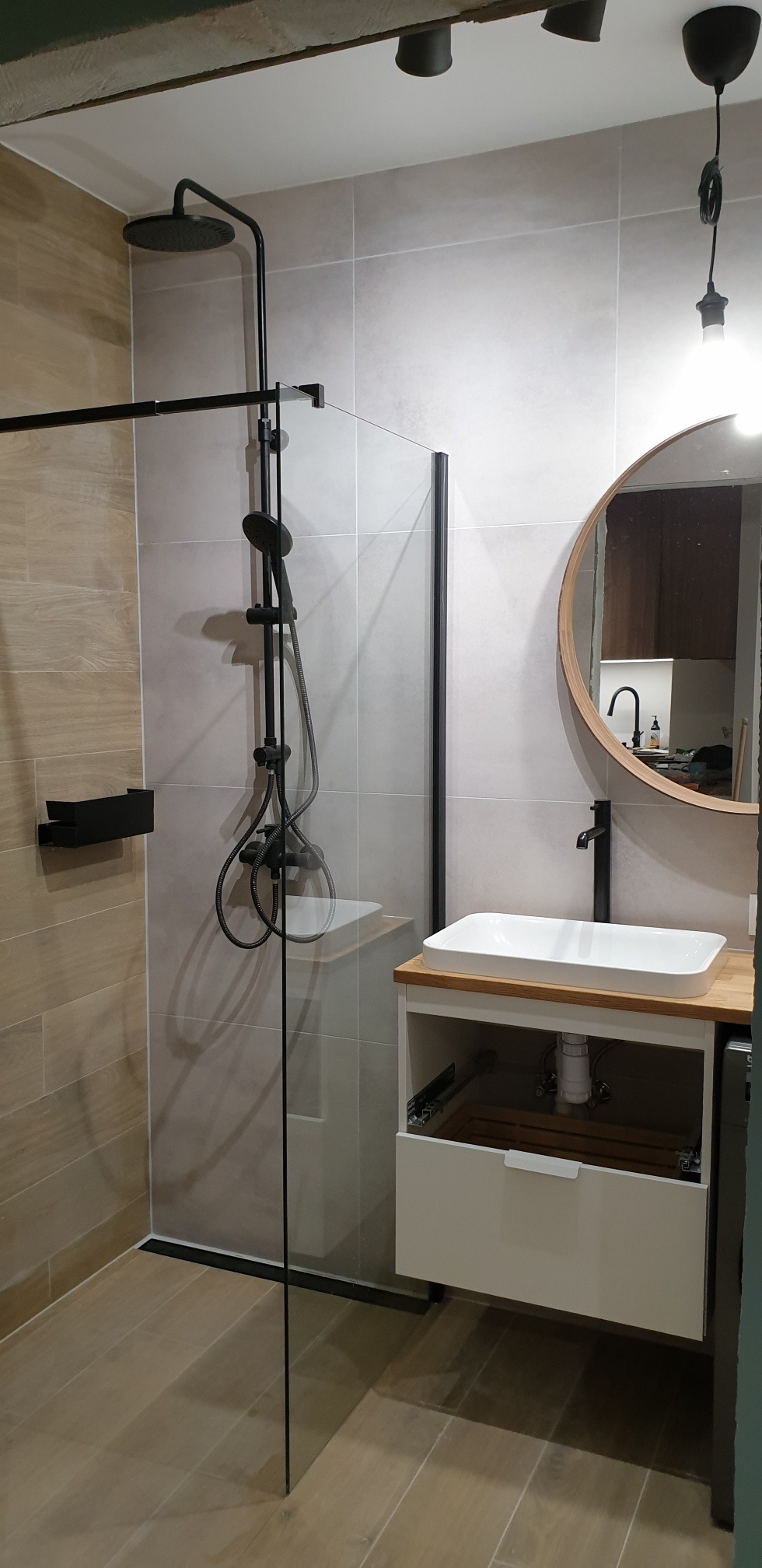 Nowoczesna łazienka z kabiną prysznicową typu walk-in z czarnymi elementami, okrągłym lustrem z drewnianą ramą i białą umywalką nablatową na szafce z drewnianym blatem.