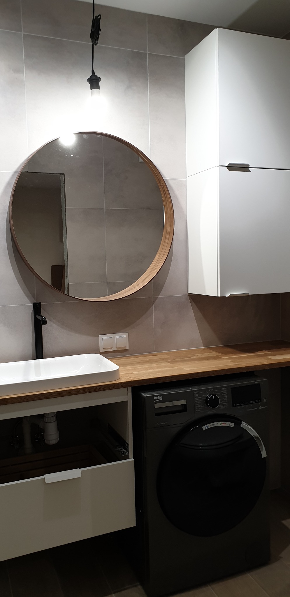 Nowoczesna łazienka z okrągłym lustrem w drewnianej ramie, białą umywalką nablatową, drewnianym blatem, zabudową meblową z pralką oraz szarymi płytkami na ścianie.