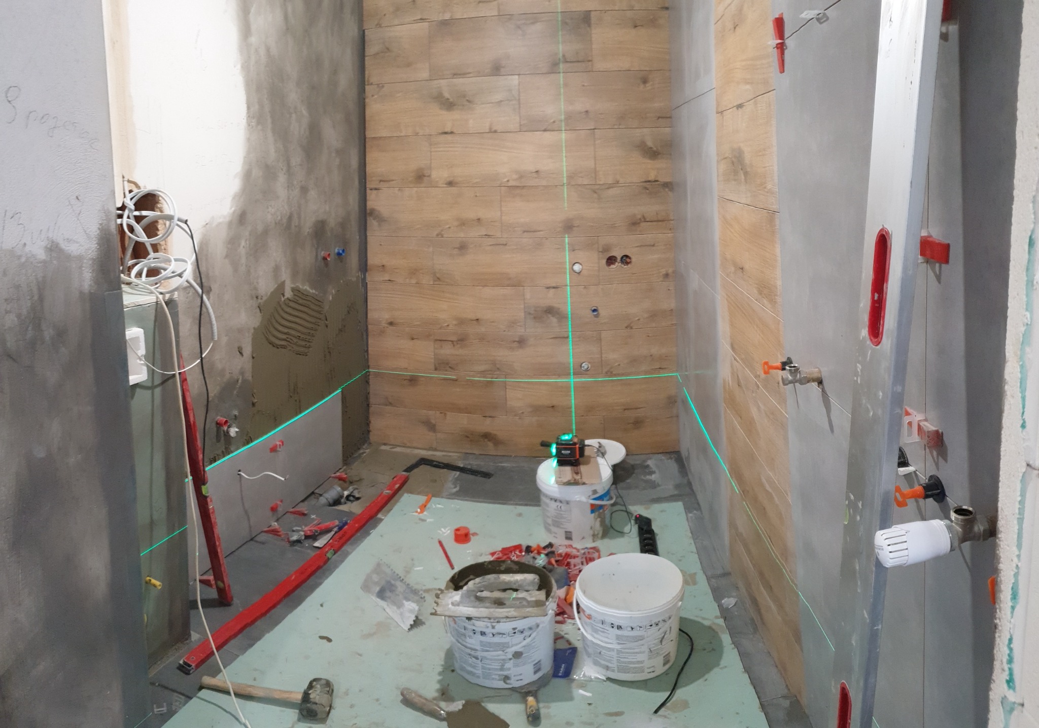 Łazienka w trakcie remontu: ściana z drewnopodobnych płytek, poziomica laserowa wyznacza linię, widoczne wiaderka z zaprawą, poziomica i młotek