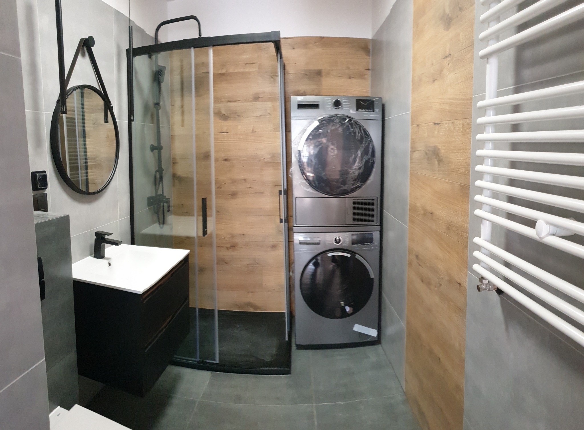 Nowoczesna łazienka z kabiną prysznicową typu walk-in, umywalką z szafką w kolorze antracytowym, okrągłym lustrem na pasku oraz pralko-suszarką w zabudowie, wykończona płytkami imitującymi drewno.