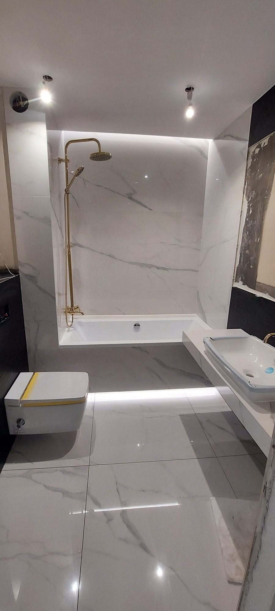 Nowoczesna łazienka z białymi marmurowymi płytkami, złotą armaturą, podświetlaną wanną i umywalką oraz wiszącą toaletą z żółtym akcentem.