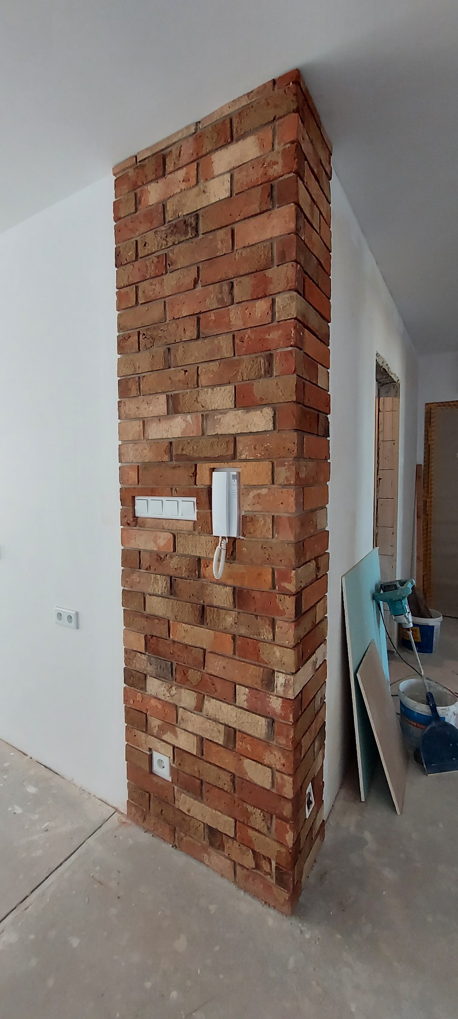 Wykończony narożnik ściany z cegły klinkierowej wewnątrz pomieszczenia, z widocznymi gniazdkami elektrycznymi i domofonem, na tle surowej podłogi i narzędzi budowlanych.