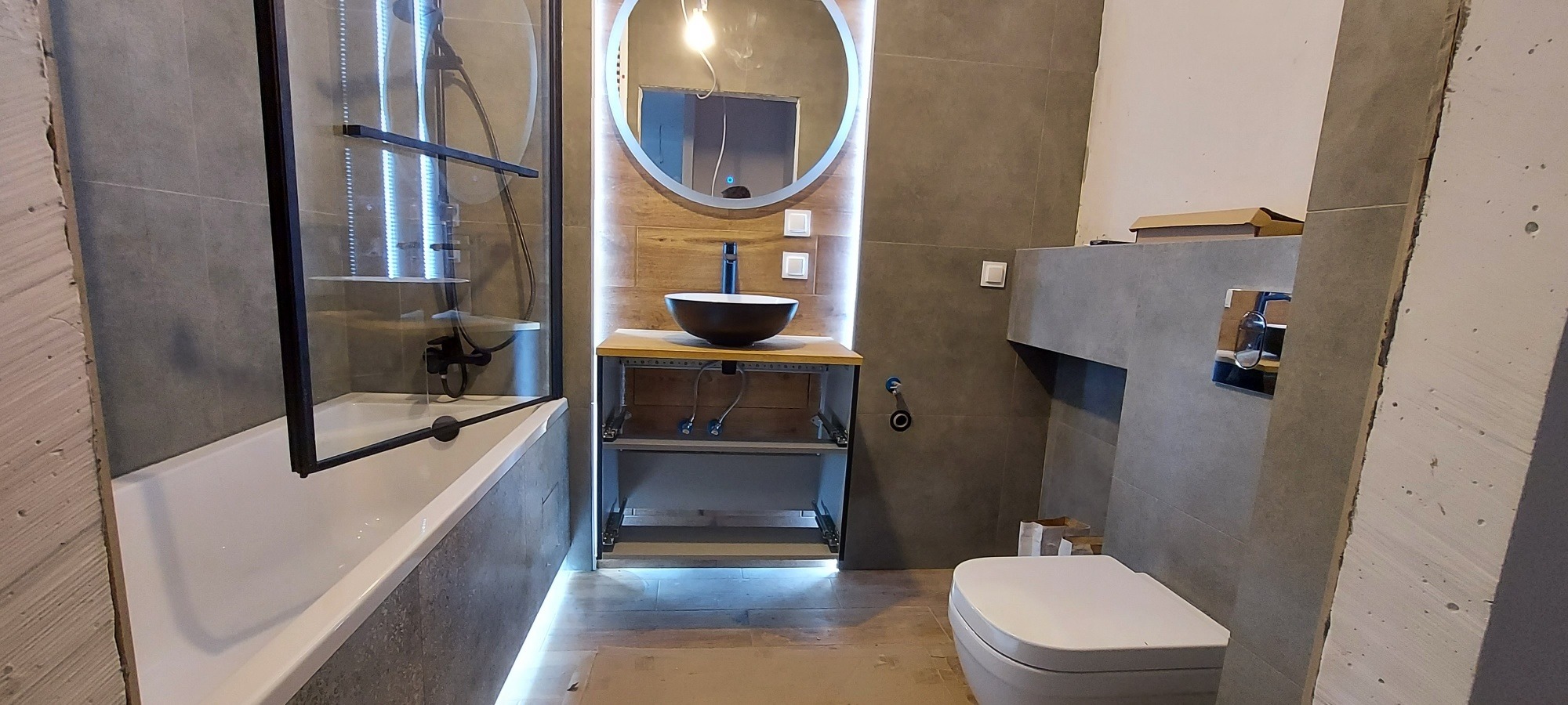 Nowoczesna łazienka w szarościach z elementami drewna, oświetlenie LED pod szafką umywalkową, czarna umywalka nablatowa i okrągłe lustro z podświetleniem, kabina prysznicowa z czarnymi ramami...