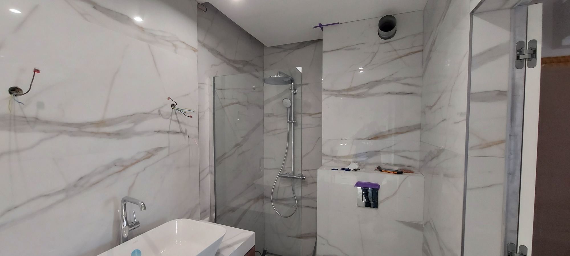 Wykończona łazienka z marmurowymi płytkami, kabiną prysznicową typu walk-in, wiszącą miską WC i umywalką nablatową. Widoczne niedokończone podłączenia elektryczne i narzędzia na półce.