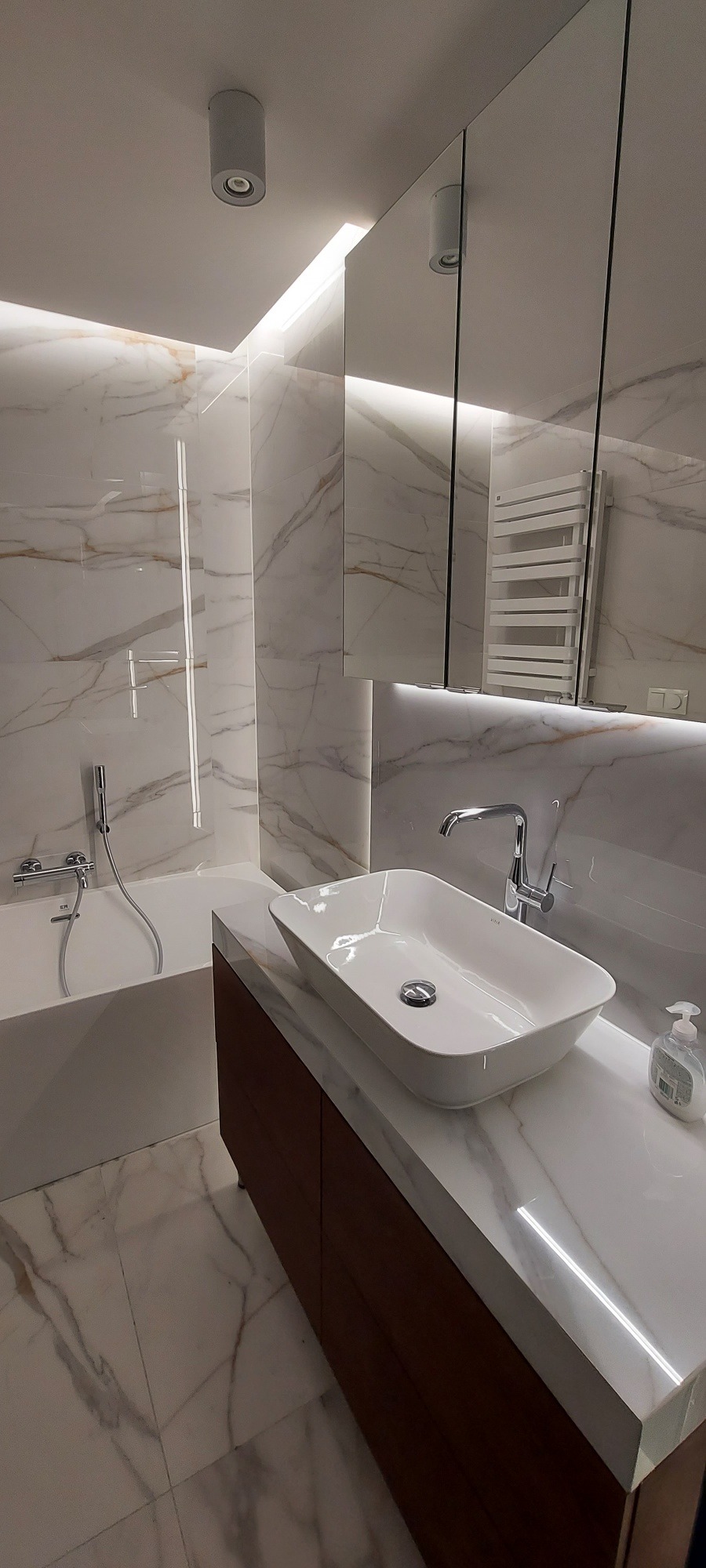 Nowoczesna łazienka z marmurowymi płytkami, podświetlanym lustrem i prostokątną umywalką nablatową, z widocznym dozownikiem mydła w płynie.