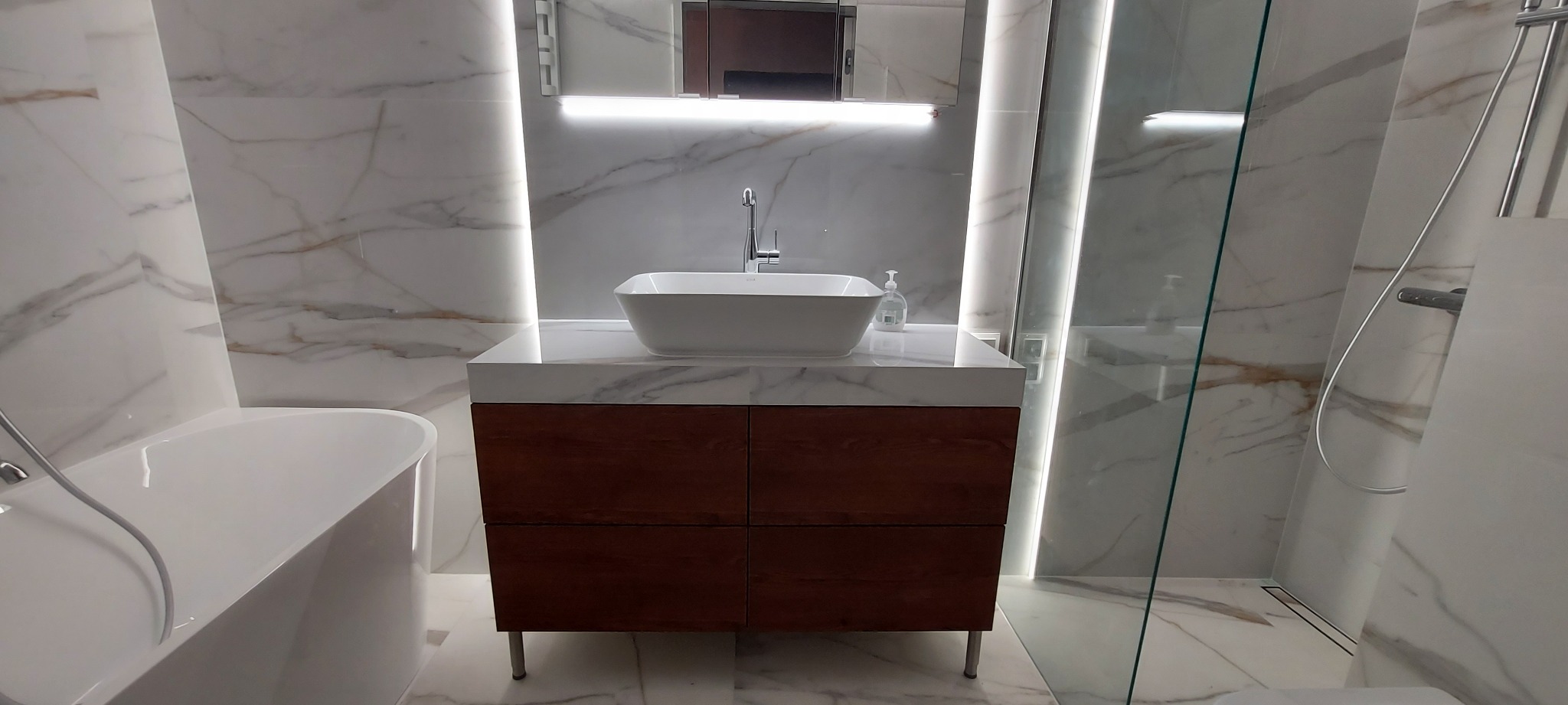 Nowoczesna łazienka z marmurowymi płytkami, drewnianą szafką pod umywalką i szklaną kabiną prysznicową, oświetlenie LED nad lustrem.