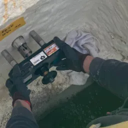 Człowiek w rękawicach trzyma urządzenie do osuszania ścian w piwnicy, widoczna żółta karteczka z datą, wilgoć na ścianach i woda w zagłębieniu