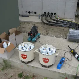 Przygotowanie do prac budowlanych: Dwa emaliowane garnki marki WEck wypełnione białymi workami z materiałem budowlanym stoją na betonowej wylewce, obok rozłożone kable elektryczne, kompresor...