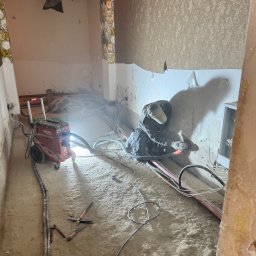 BETONTECH POLSKA SPÓŁKA Z OGRANICZONĄ ODPOWIEDZIALNOŚCIĄ - Pracownik w trakcie osuszania ściany w pomieszczeniu z dekoracyjnym sufitem, używając szlifierki kątowej i odkurzacza przemysłowego Hilti. Widoczne przewody i narzędzia.