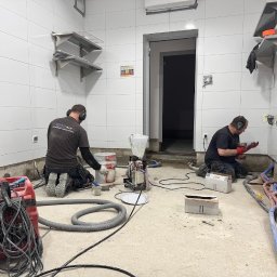 BETONTECH POLSKA SPÓŁKA Z OGRANICZONĄ ODPOWIEDZIALNOŚCIĄ - Dwóch pracowników w słuchawkach, podczas prac przygotowawczych do wylewki w pomieszczeniu z białymi płytkami. Widoczne rury, narzędzia i maszyny.