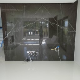 Bcs Glazura - Ściana wyłożona płytami z ciemnego marmuru z widocznymi gniazdkami elektrycznymi i uchwytem do telewizora, w trakcie montażu; przewody elektryczne leżące na jasnej podłodze.