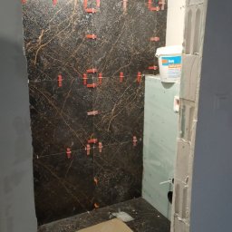 Bcs Glazura - Częściowo wyłożona ciemną, marmurkową płytką ściana łazienki z widocznymi, czerwonymi klipsami poziomującymi, jasny sufit z żarówką, fragment ściany z płyty gipsowo-kartonowej oraz wiadro z zaprawą.