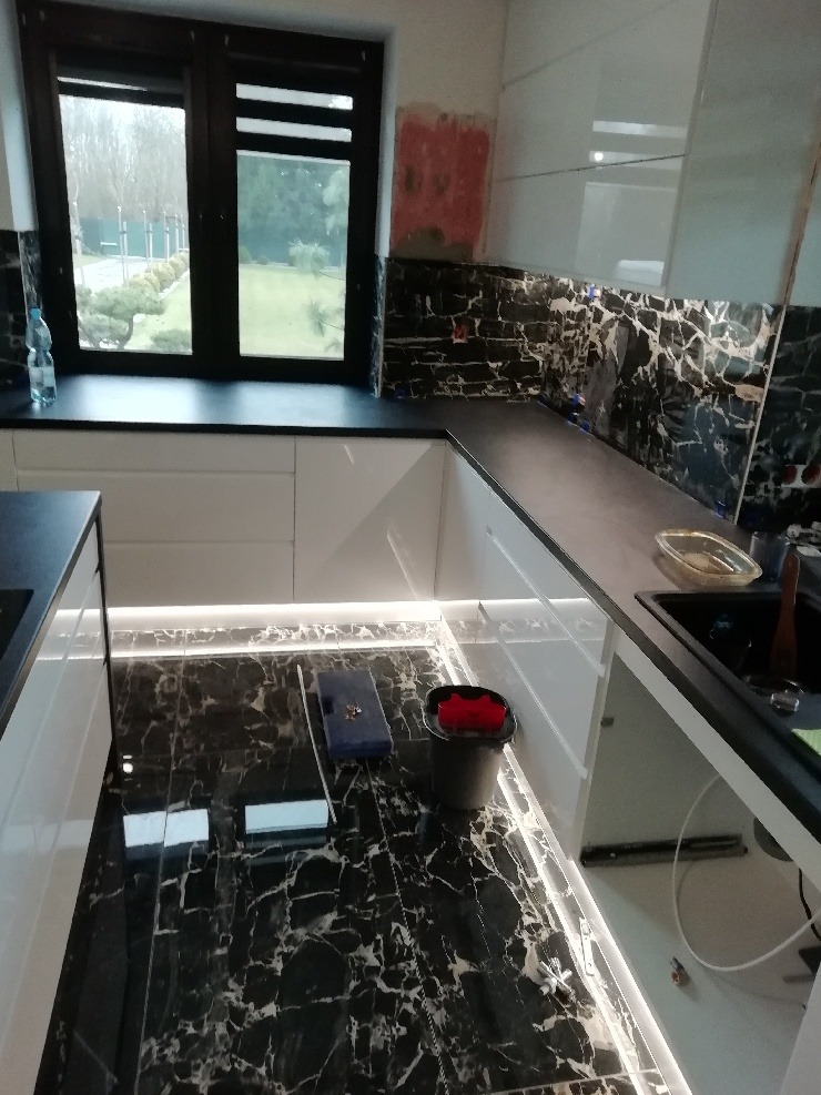 Częściowo wykończona kuchnia z czarnymi marmurowymi płytkami na podłodze i ścianie, białe szafki z podświetleniem LED, widoczne narzędzia i wiadro, fragment ściany nieotynkowany.