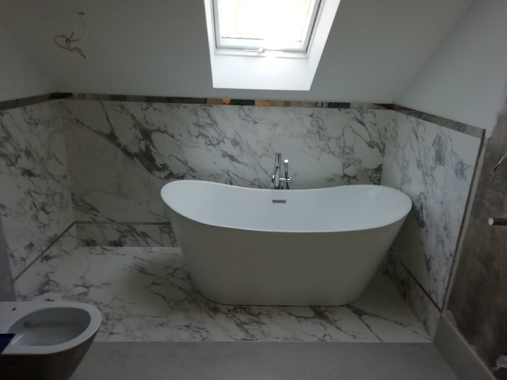 Wolnostojąca wanna na tle marmurowych płytek w łazience z oknem dachowym, widoczny fragment miski WC.