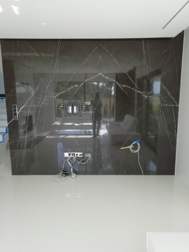 Ściana wyłożona płytami z ciemnego marmuru z widocznymi gniazdkami elektrycznymi i uchwytem do telewizora, w trakcie montażu; przewody elektryczne leżące na jasnej podłodze.