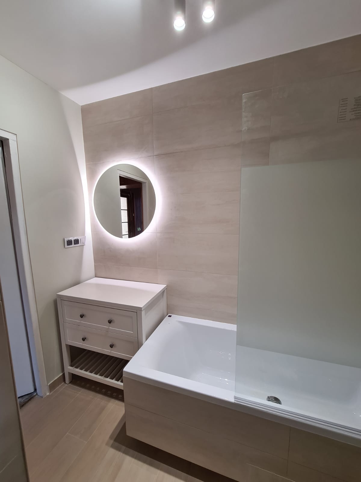 Jasna łazienka po remoncie: beżowe płytki, biała wanna z parawanem, szafka z umywalką i okrągłe lustro z podświetleniem LED.