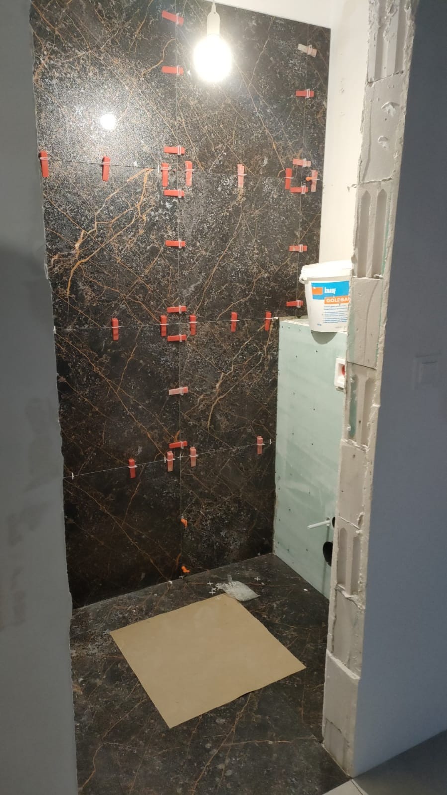 Częściowo wyłożona ciemną, marmurkową płytką ściana łazienki z widocznymi, czerwonymi klipsami poziomującymi, jasny sufit z żarówką, fragment ściany z płyty gipsowo-kartonowej oraz wiadro z zaprawą.