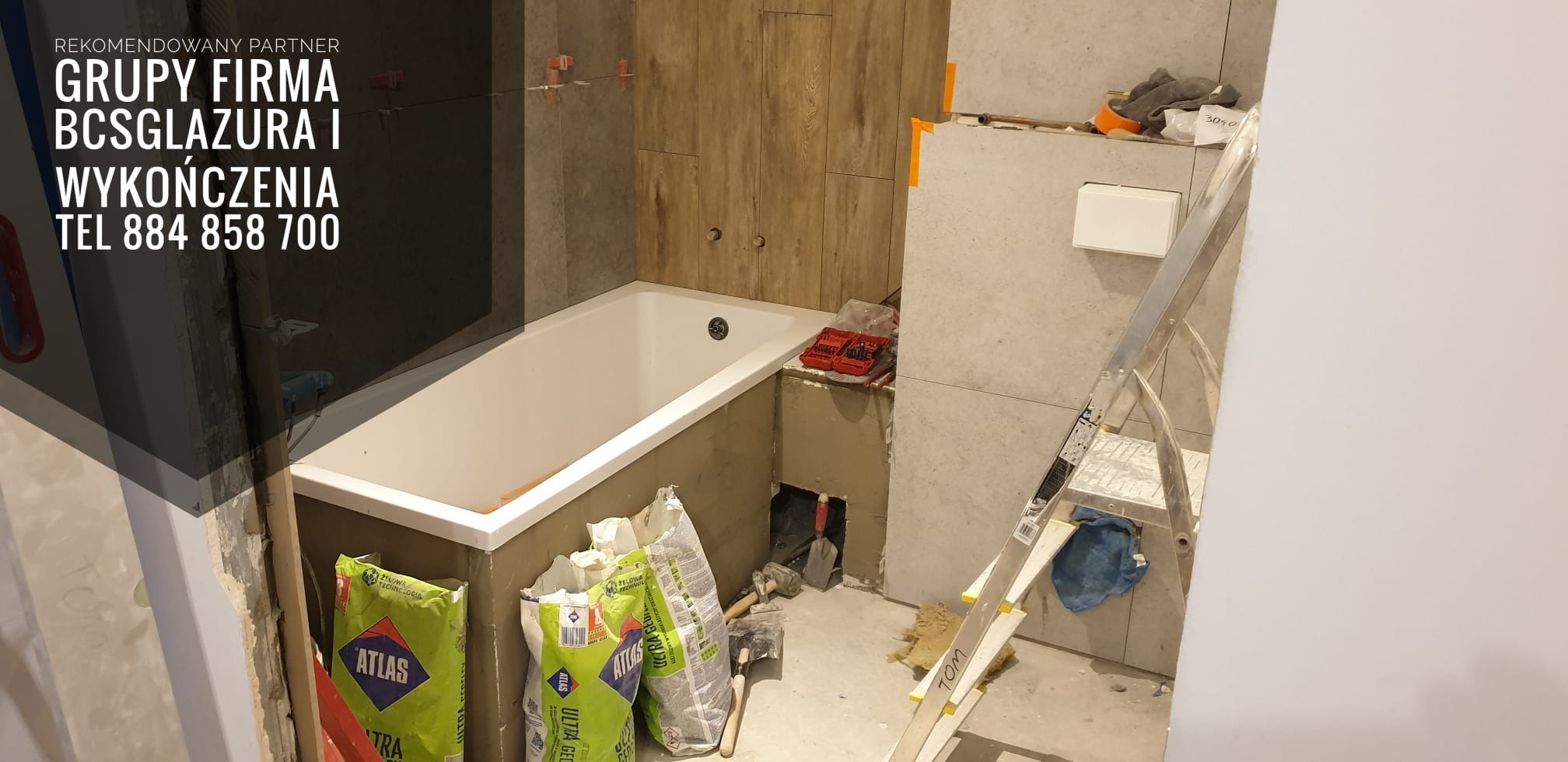 Łazienka w trakcie prac wykończeniowych: wanna obudowana płytkami imitującymi drewno, częściowo wyłożone ściany, drabina oparta o ścianę, worki z zaprawą Atlas na podłodze, widoczne narzędzia...