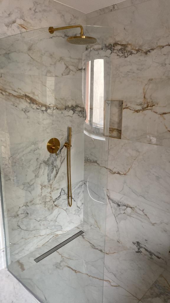 Nowoczesny prysznic typu walk-in z marmurowymi płytami, złotą armaturą i odpływem liniowym.