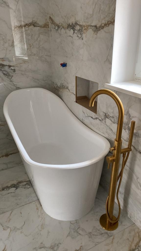 Wolnostojąca wanna na tle marmurowych płytek, z złotą baterią podłogową i wnęką w ścianie, prezentująca eleganckie wykończenie.