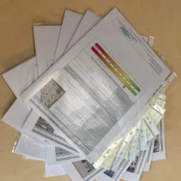 Stos świadectw charakterystyki energetycznej budynków ułożonych na drewnianym blacie, widoczne wykresy i tabele z danymi, fragmenty zdjęć budynków, ponumerowane przekładki.