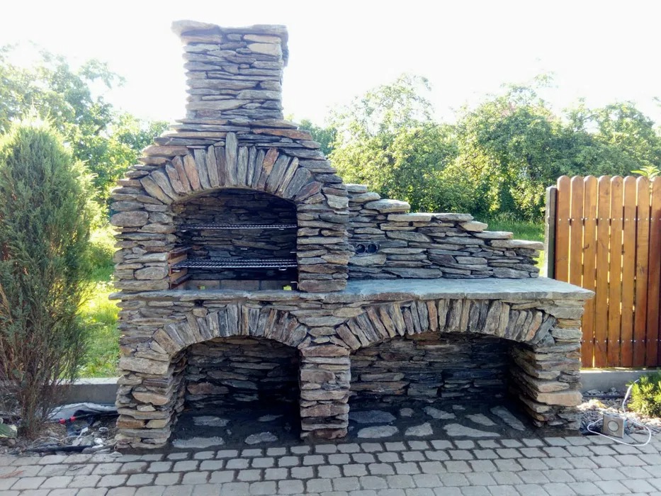 Kamienny grill ogrodowy z wędzarnią, zbudowany z nieregularnych płaskich kamieni, z widocznym rusztem wewnątrz paleniska i dwoma wnękami na drewno pod spodem, na tle zieleni i drewnianego płotu.