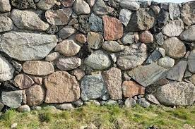 Masywny mur z nieregularnych, różnobarwnych kamieni polnych, z widocznymi fragmentami trawy u podstawy, tworzący rustykalną teksturę.