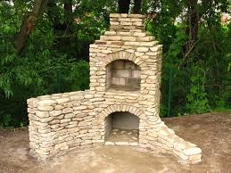 Murowany grill ogrodowy z jasnego kamienia, z dwoma łukowymi otworami, na tle zieleni drzew.