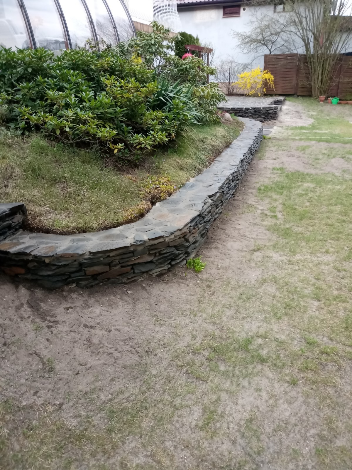 Kamienna ścianka oporowa oddzielająca rabatę z roślinami od trawnika, wykonana z nieregularnych, płaskich kamieni, z widoczną ścieżką z kamiennych płyt w tle.