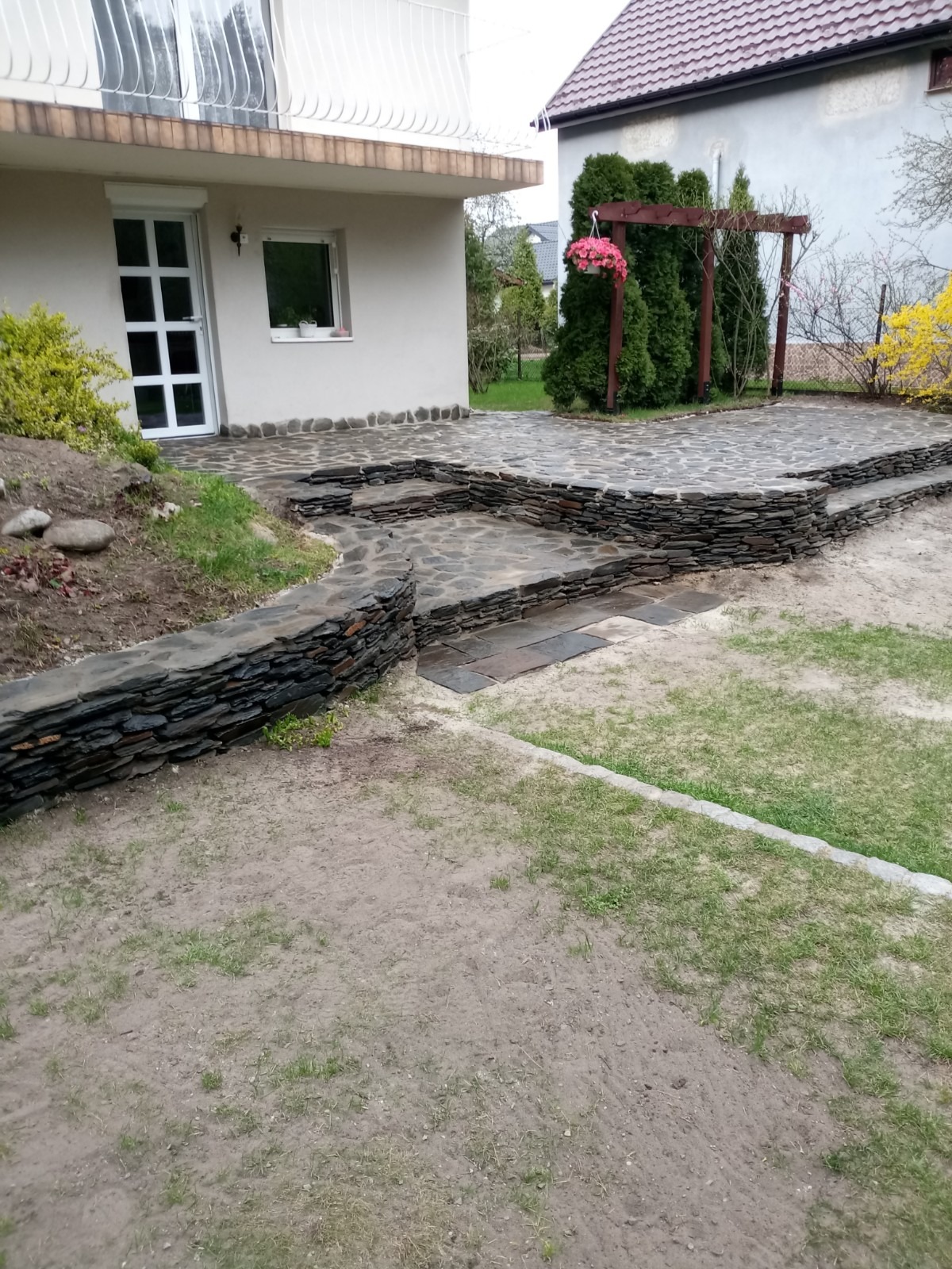 Kamienny taras z nieregularnych płyt, schodkowy układ, widok na dom z białymi drzwiami i drewnianą pergolą z wiszącymi kwiatami.
