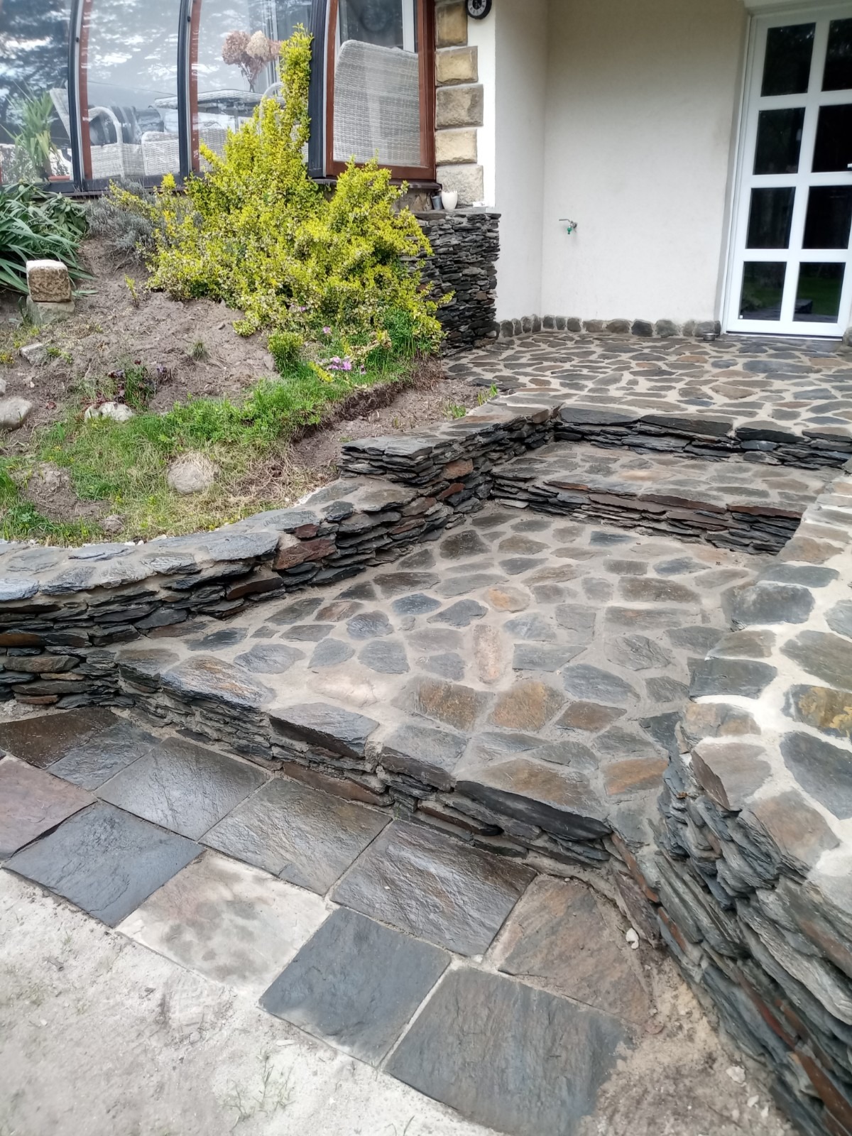 Kamienny taras z nieregularnych płyt i schodów, wykończony obrzeżem z łupka, prowadzący do wejścia budynku z białymi drzwiami.