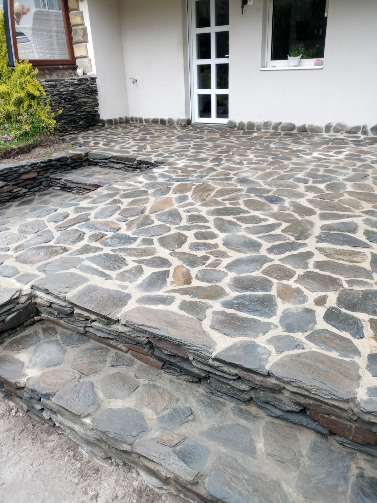 Kamienny taras z nieregularnych płyt, schody wykończone kamieniem, jasna elewacja budynku w tle.