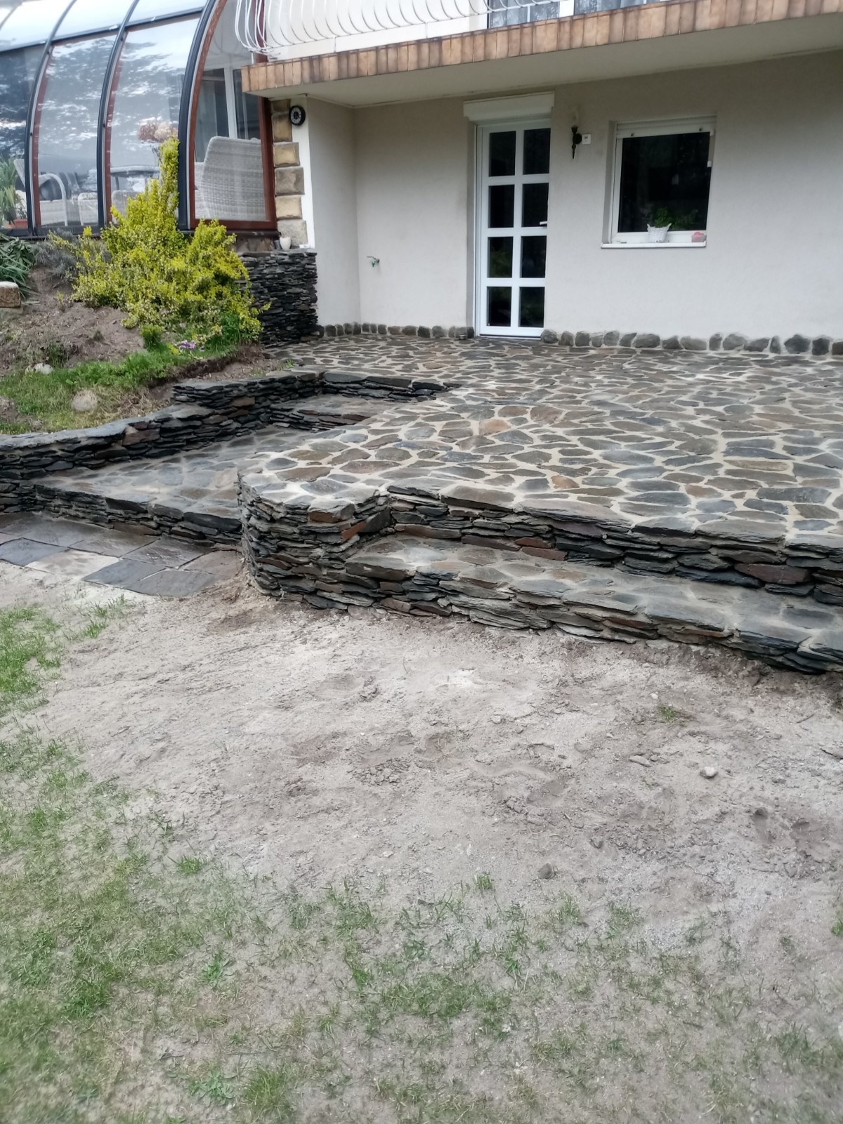 Kamienny taras z nieregularnych płyt, prowadzący do domu z przeszkloną werandą i białymi drzwiami, wykończony warstwami kamienia łupkowego.