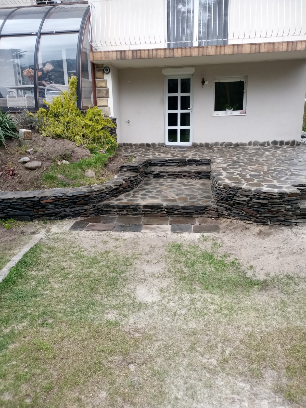 Kamienny taras przed domem z mozaikową nawierzchnią i schodami wykończonymi łupkiem, prowadzącymi do białych drzwi.