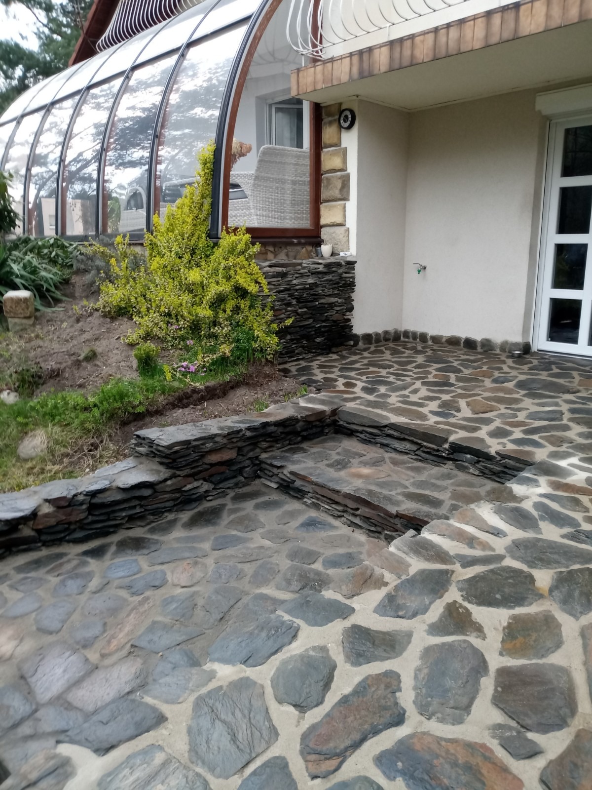 Kamienny taras z nieregularnych płyt, schodki z łupka, widok na oranżerię i fragment elewacji domu.