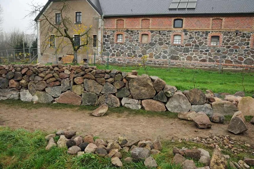 Kamienny murek z nieregularnych głazów w rustykalnym stylu, oddzielający trawnik od ścieżki, na tle budynku z elewacją częściowo kamienną i ceglaną.