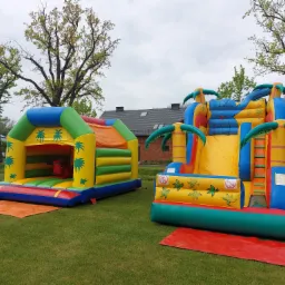 Dwa kolorowe, dmuchane zamki na trawie, jeden z motywem dżungli z palmami, drugi w kształcie domku z czerwonym bykiem w środku, na tle drzew i budynku.