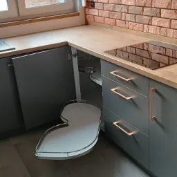 Szara zabudowa kuchenna z drewnianym blatem i ceglaną ścianą w tle, widoczny otwarty narożny system półek obrotowych.